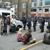 Riot cop leans on CBC vehicle while peaceful protestors sit by