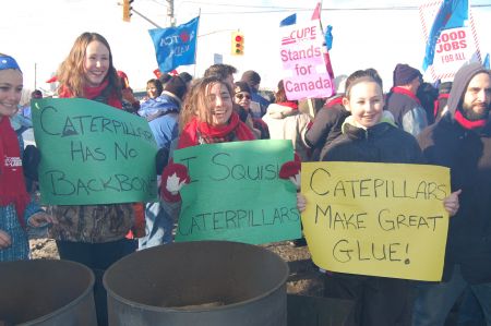 Three young women hold playful protest signs outside the front gate of Electro-Motive Diesel during a mass picket in London, Ont. on Jan. 21  Photo: Mick Sweetman 